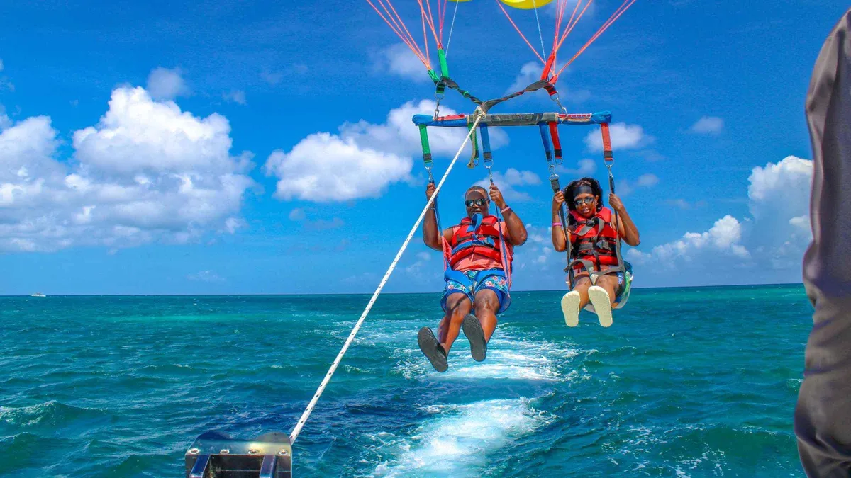 Two people parasailing high above turquoise water off Palm Beach with a photographer on the boat below, Aruba